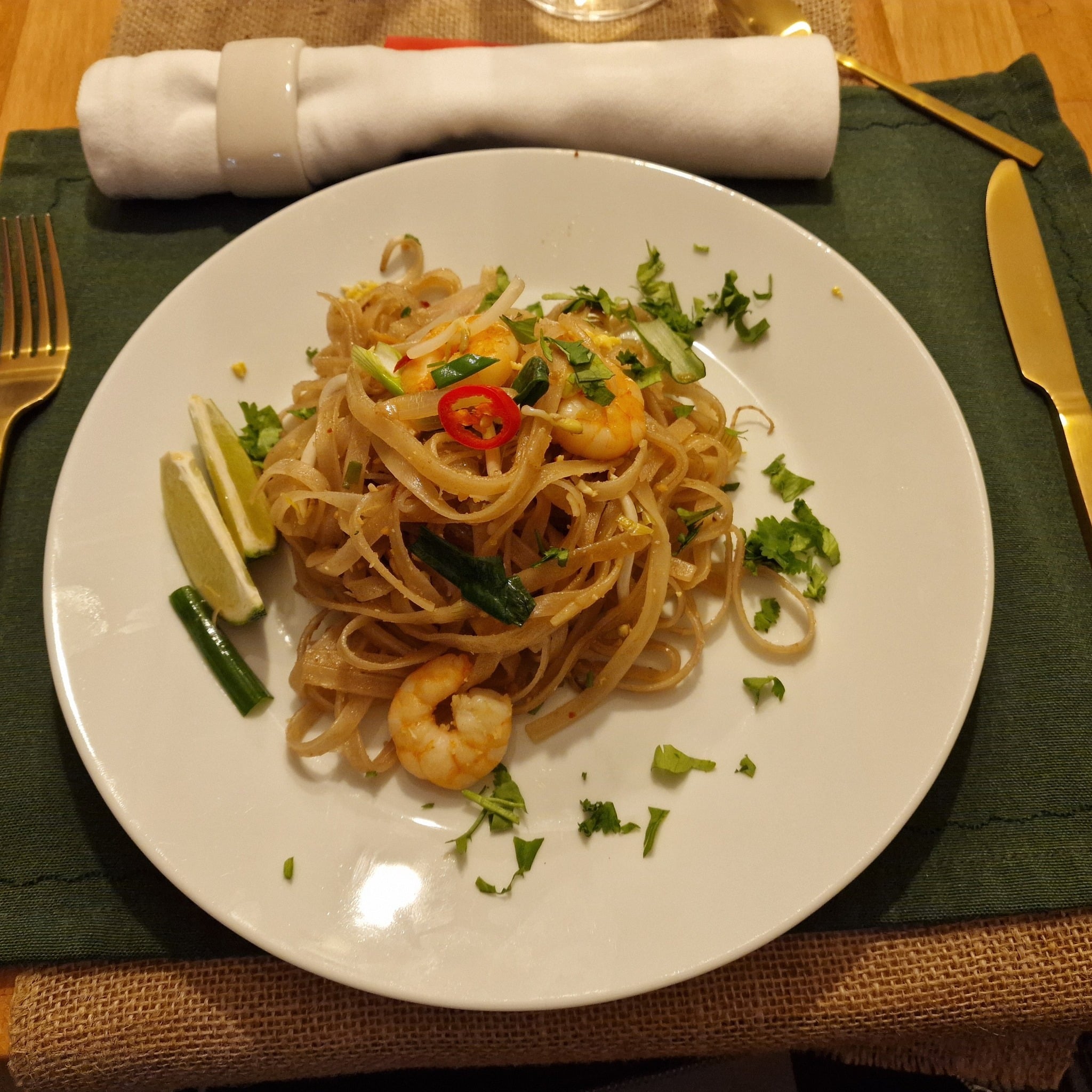 Ein Teller Pad Thai mit Garnelen, garniert mit Koriander, Chilischeiben und Frühlingszwiebeln. Limettenspalten liegen dazu. Zum Tischgedeck gehören eine ordentlich gerollte weiße Serviette und goldenes Besteck.
