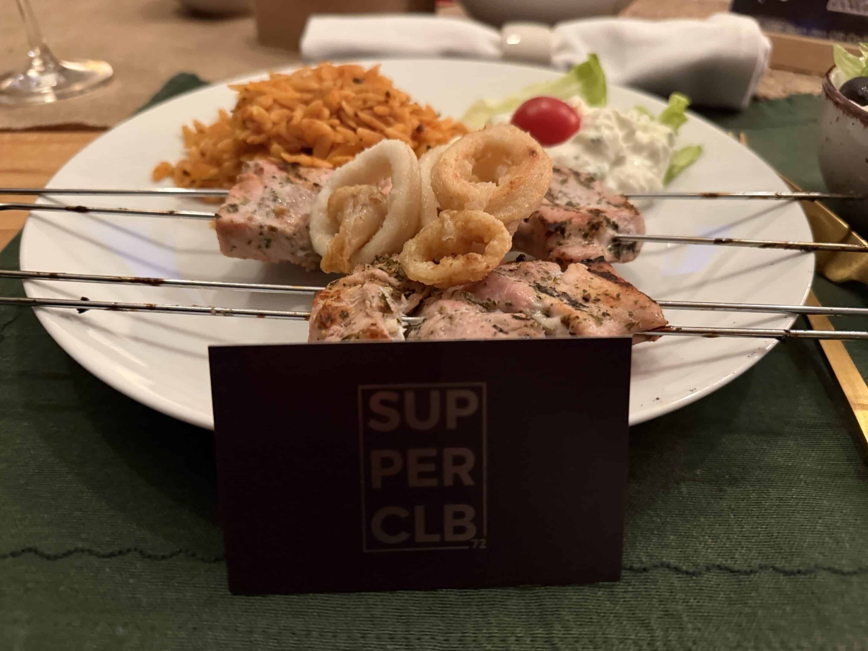 Ein Teller mit Spießen aus gegrilltem Fleisch und frittierten Calamari, serviert mit Reis und einem Klecks Tzatziki-Sauce. Auf der grünen Tischdecke im Vordergrund liegt eine Karte mit SUPER CLB.