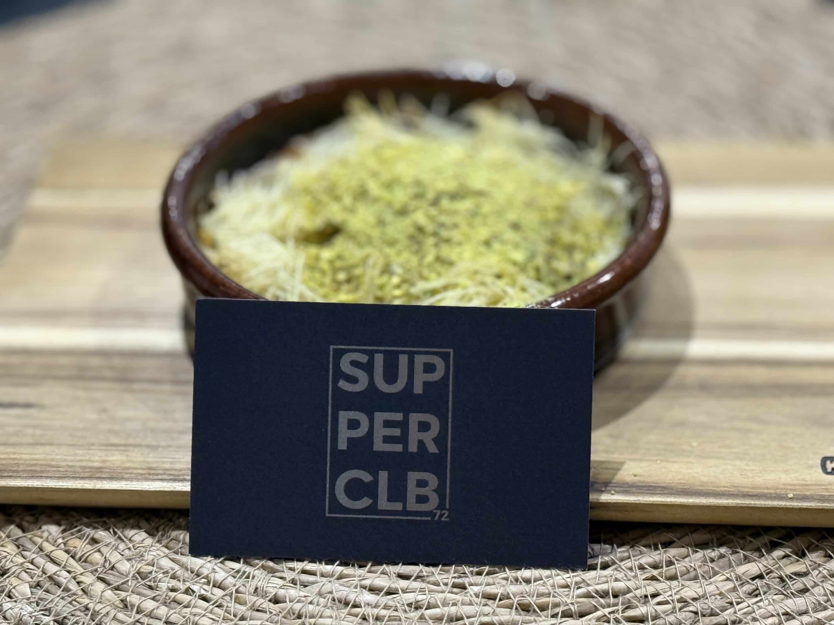 Ein Gericht mit zerkleinertem Teig und zerstoßenen Pistazien steht in einer braunen Schüssel auf einem Holzbrett. Vor der Schüssel steht auf einer dunklen Karte in fetten, hellen Buchstaben „SUPER CLB. 72“.