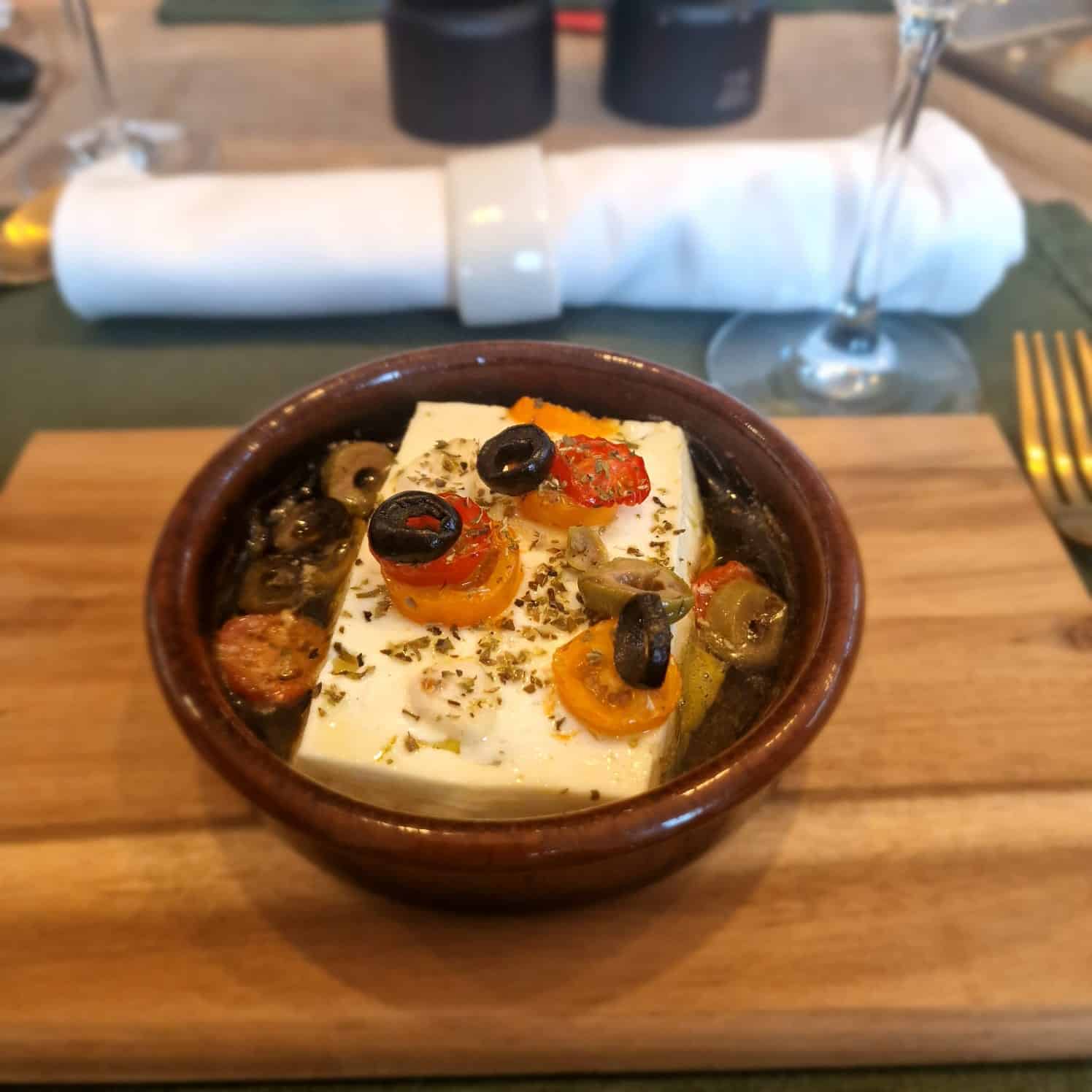 Auf einer Keramikschale liegt gebackener Fetakäse mit Kirschtomaten, schwarzen Oliven und Kräutern. Serviert wird er auf einem Holzbrett, im Hintergrund steht ein Glas Champagner, daneben eine ordentlich gefaltete Serviette, Besteck und eine Flasche.