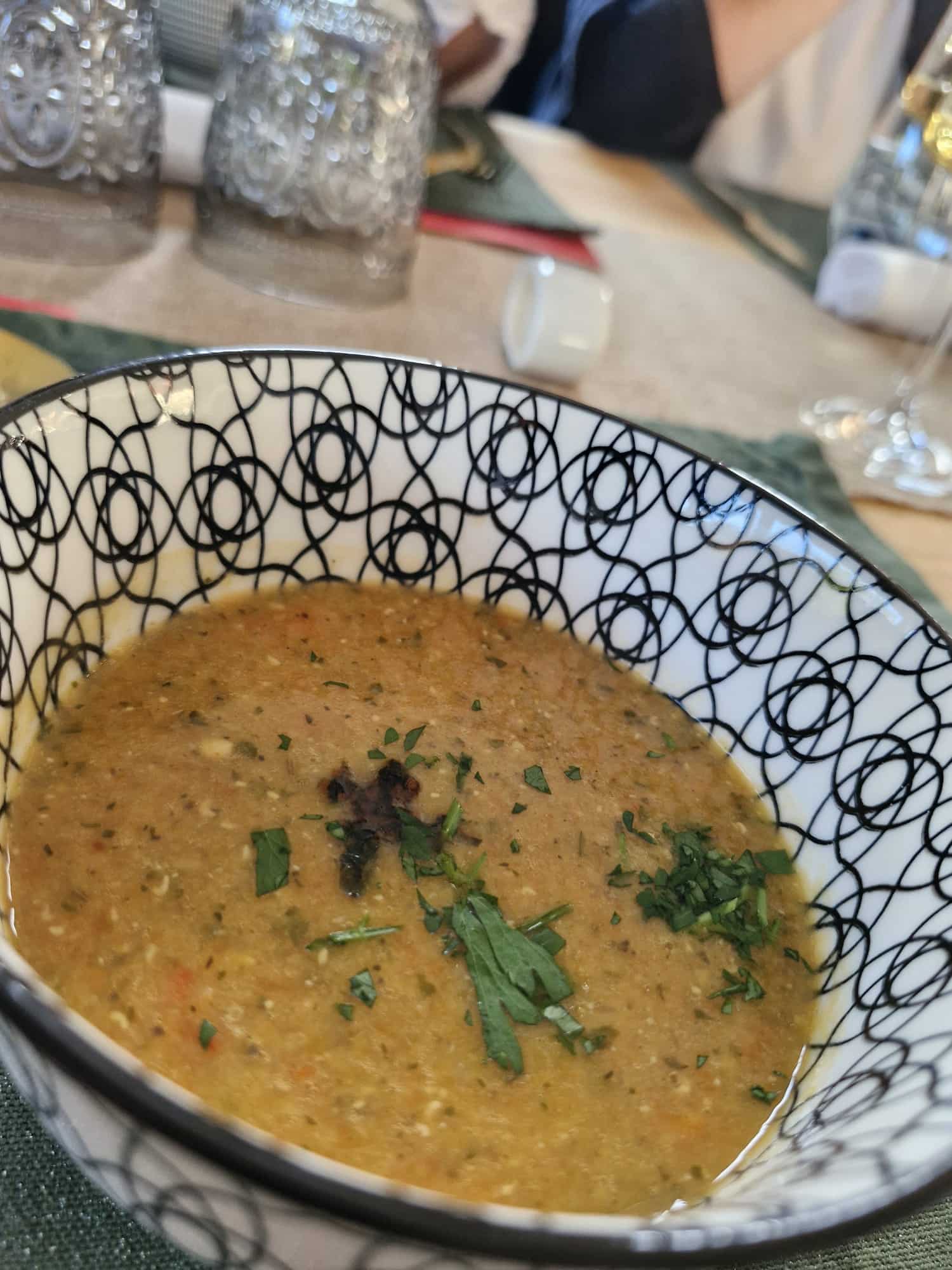 Auf einem Tisch steht eine gemusterte Schüssel mit einer dicken, gelbbraunen Suppe, garniert mit gehackten Kräutern. Im Hintergrund sind Glasgeschirr und andere Essgeschirre unscharf zu sehen.