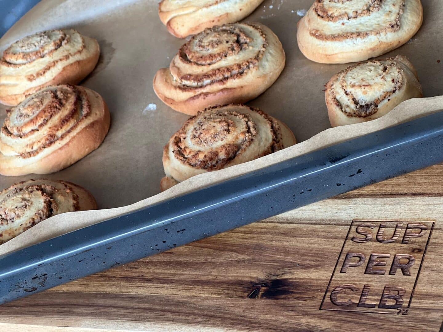 Ein Tablett mit frisch gebackenen Zimtbrötchen liegt auf einem mit Pergament ausgelegten Backblech, das teilweise auf einem Holzbrett mit der Aufschrift SUPPER CLUB.72 ruht.