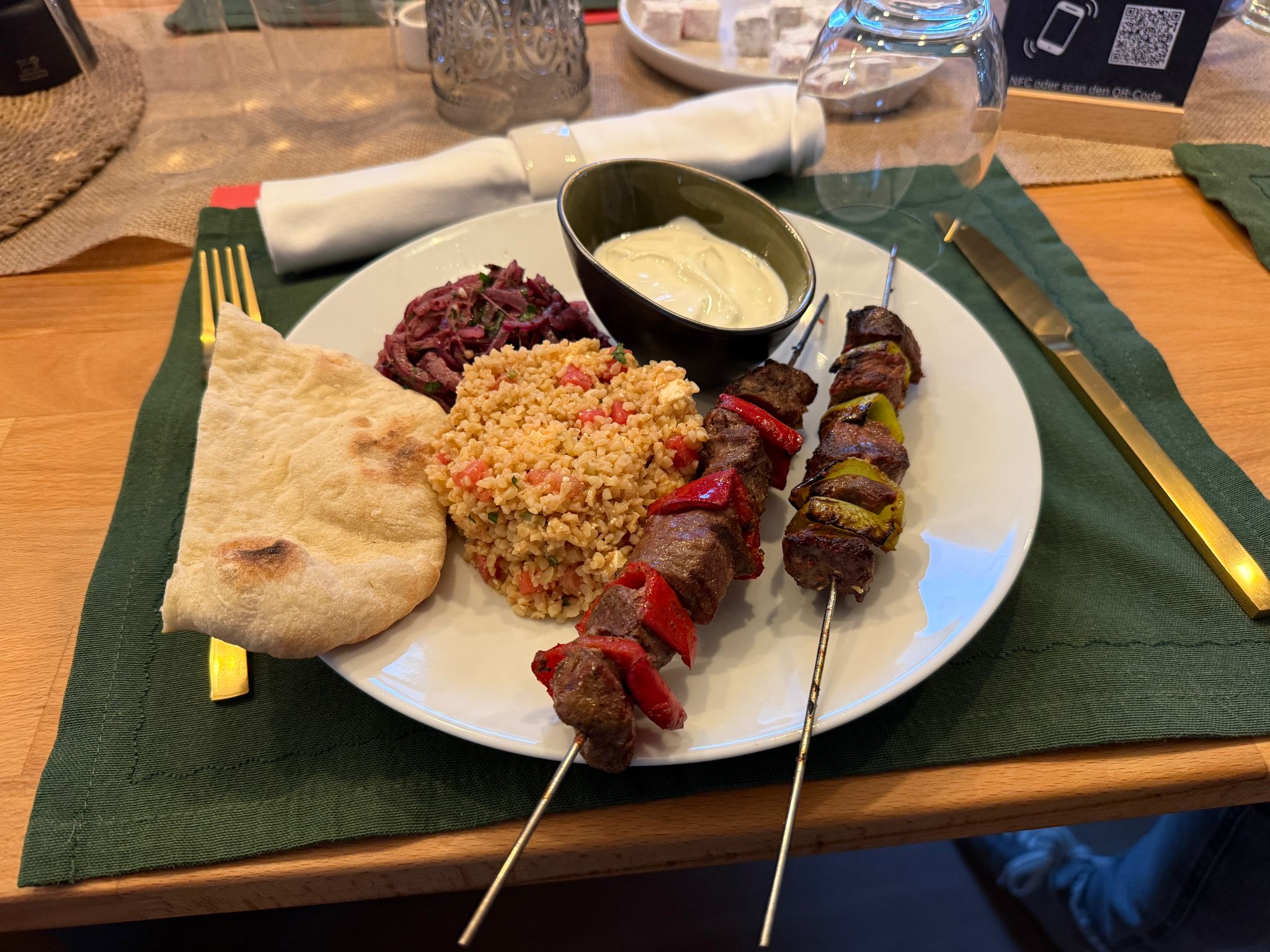 Ein Teller mit zwei Rindfleischspießen, gegrilltem Gemüse, Couscous, einem Fladenbrot, Rotkohlsalat und einer Schüssel weißer Soße, auf einem grünen Tischset mit goldfarbenem Besteck und einem Glas.