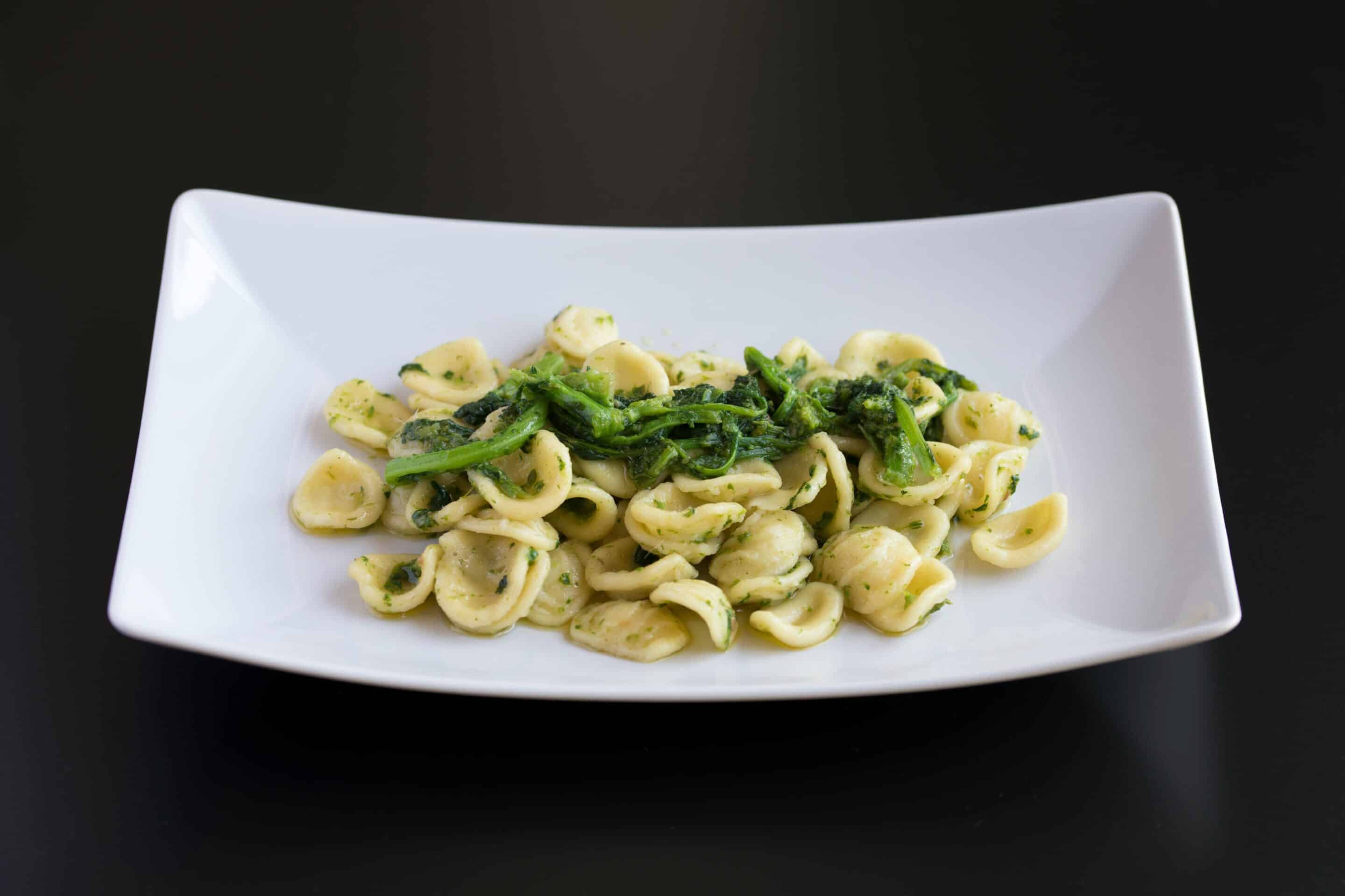 Ein weißer rechteckiger Teller mit Orecchiette-Nudeln, belegt mit grünem Blattgemüse, wahrscheinlich Stängelkohl, in einer hellen, grünen Kräutersauce geschwenkt, vor einem dunklen Hintergrund.