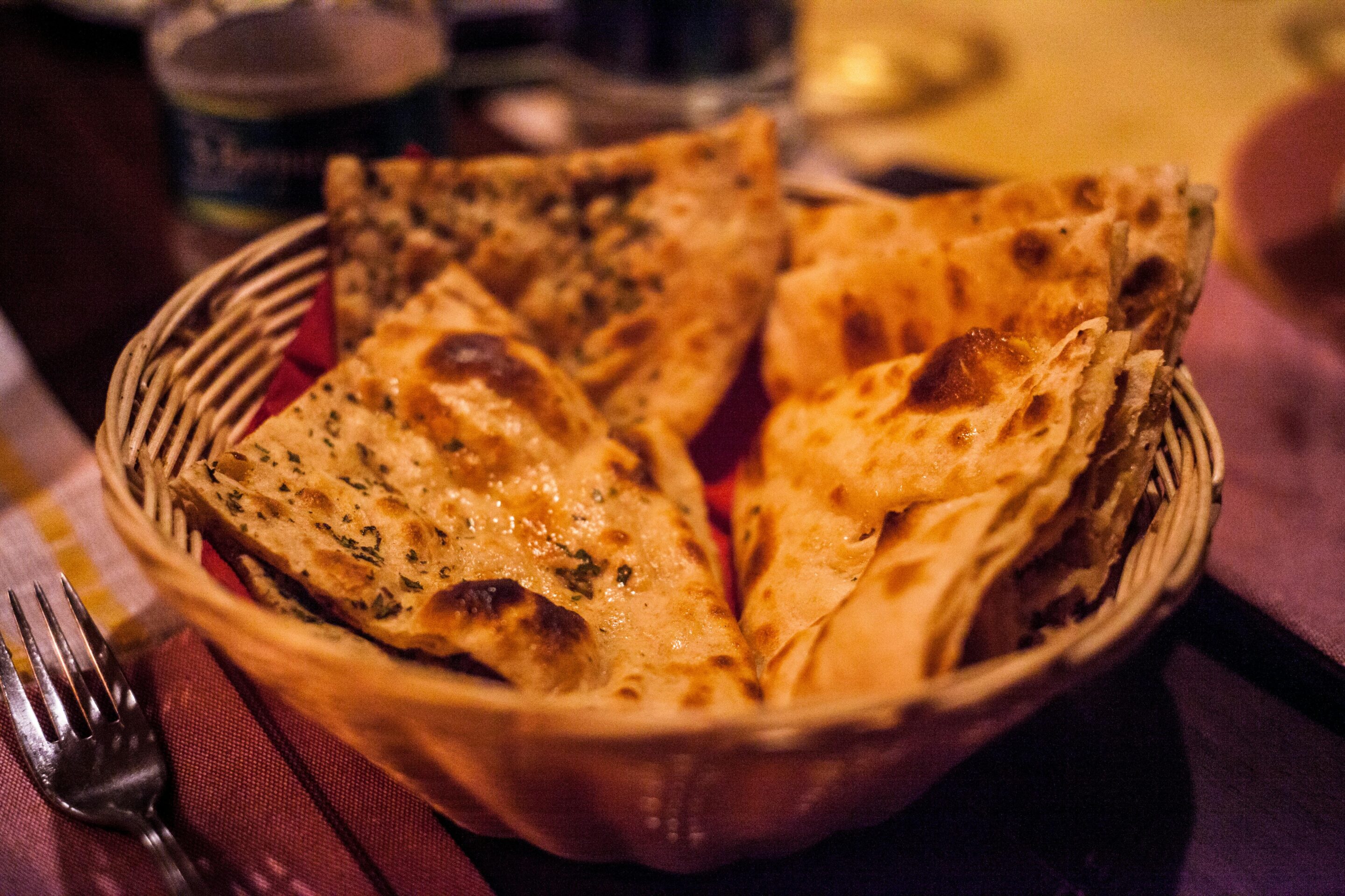 Ein Korb gefüllt mit verschiedenen Naan-Brotstücken, einige davon mit Kräutern und Gewürzen garniert, steht auf einem für eine Mahlzeit gedeckten Tisch.