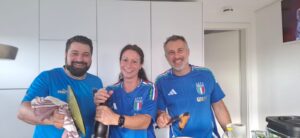 Drei lächelnde Menschen in blauen Italien-Fußballtrikots stehen in einer Küche, halten Küchenutensilien und Lebensmittel in der Hand und scheinen gemeinsam zu kochen. Im Hintergrund sind weiße Schränke und ein Fernseher an der Wand zu sehen.