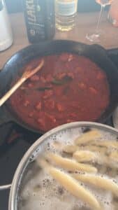 Eine Pfanne mit Tomatensoße und Fleischstücken wird mit einem Holzlöffel neben einem Topf mit kochenden Nudeln auf einer Herdplatte umgerührt. Im Hintergrund sind Flaschen mit Öl und ein Weinglas zu sehen.