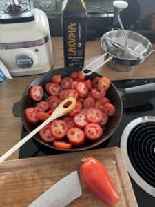Auf der Herdplatte steht eine gusseiserne Pfanne mit geschnittenen Tomaten und einem Holzlöffel. In der Nähe liegen ein Schneidebrett mit einem Messer und einer ganzen Tomate, eine Flasche Olivenöl, eine Küchenmühle und ein Standmixer.