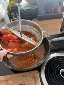 Eine Hand presst geschnittene Tomaten durch eine Mühle über einem Topf auf dem Herd, um Tomatensauce herzustellen. In der Nähe sind eine Flasche Olivenöl und ein Schneidebrett zu sehen.