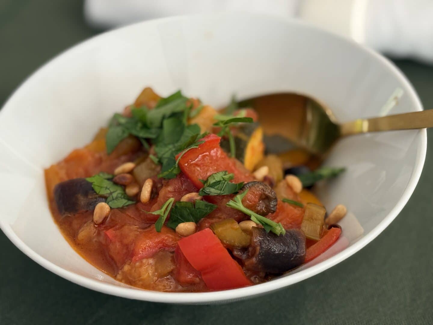 Eine weiße Schale, gefüllt mit farbenfrohem Ratatouille, garniert mit frischen Kräutern und Pinienkernen, in der ein goldener Löffel auf einer grünen Fläche ruht.