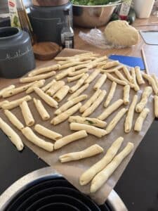 Ungebackene Stangen aus gerolltem Teig liegen auf Pergamentpapier auf einem Küchentisch. In der Nähe stehen Küchenbehälter, eine Flasche Öl, eine Teigkugel und eine Schüssel mit Grünzeug.