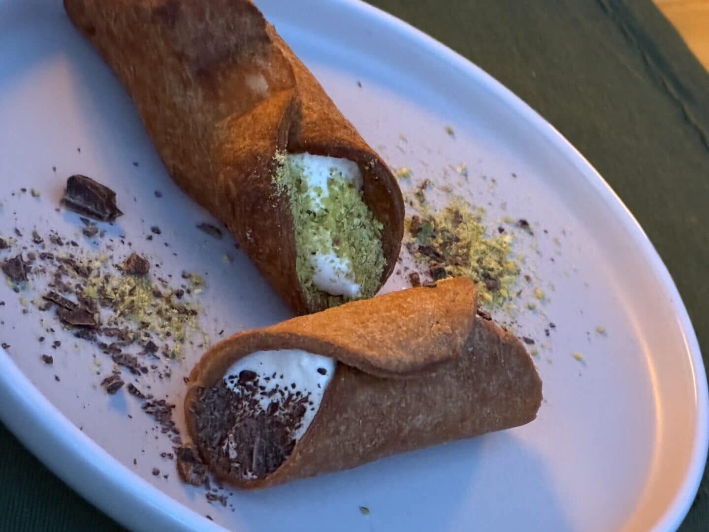 Cannoli Siciliani Zwei mit Sahne gefüllte Cannoli, die mit gehackten Pistazien und Schokoladenflocken bestreut sind, liegen auf einem weißen ovalen Teller, um den herum noch mehr Schokolade und Pistazien verstreut sind.
