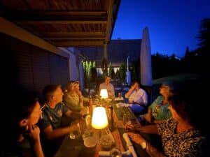Eine Gruppe von sieben Personen sitzt in der Abenddämmerung um einen Tisch im Freien, der von warmem Lampenlicht erhellt wird. Sie unterhalten sich, lachen und genießen Getränke und Snacks in einem gemütlichen Hinterhof. Im Hintergrund sind Bäume und Gebäude zu sehen.