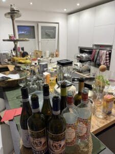Eine überfüllte Küchentheke mit leeren Wein- und Schnapsflaschen, Gläsern, einem Mixer, Obst, Geschirr und verschiedenen Lebensmitteln, die verstreut herumliegen und auf eine kürzliche Party oder ein Treffen in einer modernen Küche hindeuten.