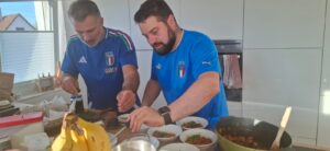 Zwei Männer in blauen italienischen Fußballtrikots bereiten in einer hellen, modernen Küche Speisen zu. Auf dem Tresen stehen Schüsseln mit Lebensmitteln und im Vordergrund Bananen. Durch ein Fenster strömt Sonnenlicht herein.